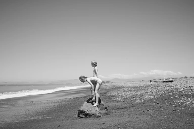 Two children standing on a large rock on a pebble beach near the ocean under a clear sky.