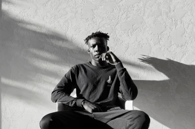 Monochrome portrait of a young Black man seated outdoors against a textured wall with strong harsh shadows.