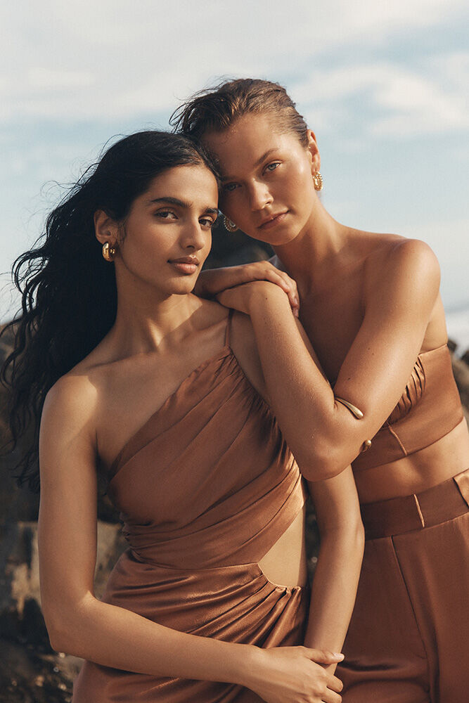 Two women in brown dresses pose closely against a rocky, outdoor backdrop with a glimpse of the ocean and sky.