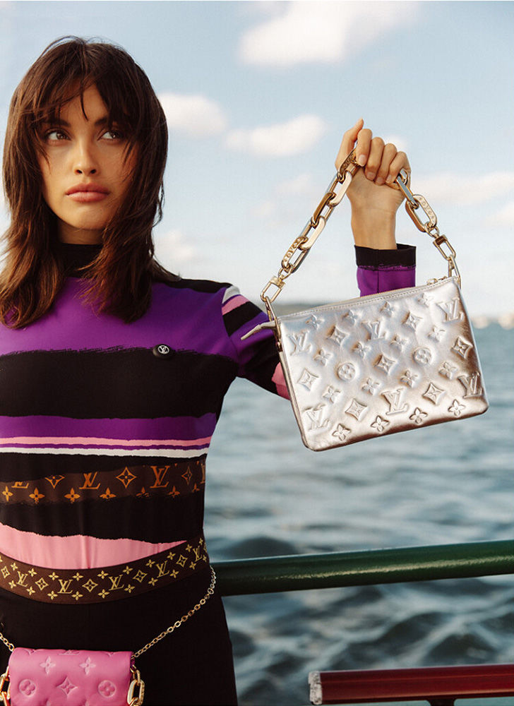 Woman posing with a silver embossed handbag in front of blue water and sky, wearing a colorful top and pink purse.