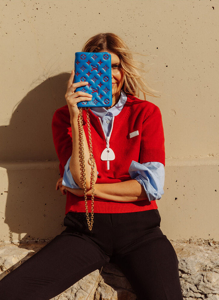 Woman with blonde hair sitting outdoors and holding a blue patterned purse with a gold chain over her face, smiling in sunlight.