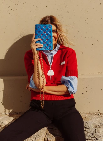 Woman with blonde hair sitting outdoors and holding a blue patterned purse with a gold chain over her face, smiling in sunlight.