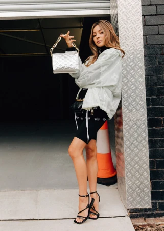 Woman in white jacket and black shorts leaning against a brick wall holding a silver handbag in Sydney