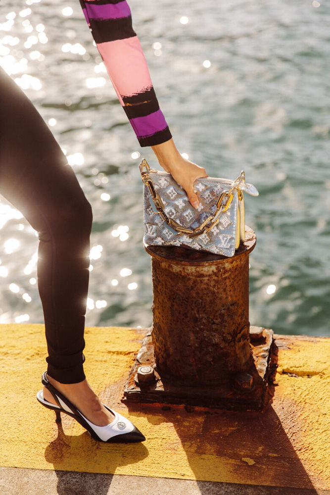 Model showcasing Louis Vuitton purse and slingback heels while posing on a yellow and rust coloured pier with water backdrop.