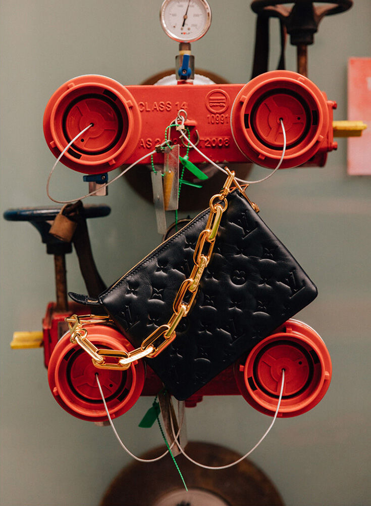 Black Louis Vuitton purse with a gold chain hangs on industrial equipment with a pressure gauge and red spigot valves indoors.