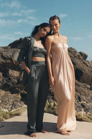Two women model satin outfits on a beach, one in a strapless peach gown and the other in a green blazer and pants set, against a blue sky.