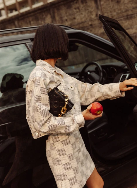 A woman in a checkered jacket and skirt stands by a black SUV, holding a black purse with a gold chain and a red apple.