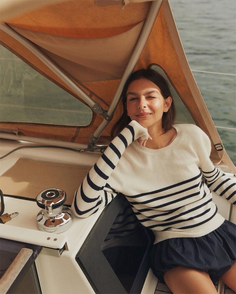 A young woman in a striped shirt sits on a sailboat with white sails, enjoying a sunny day out on the water, captured in a portrait.