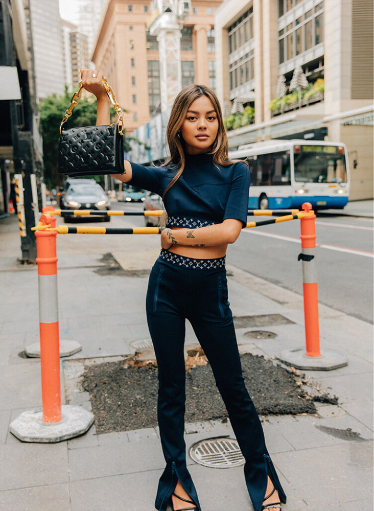 Brunette woman poses with a black handbag in an urban setting, wearing a two-piece navy blue outfit with high heels on a city street.