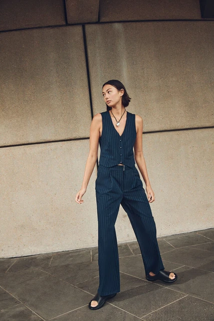 Woman poses in a pinstripe vest and wide-leg trousers against a textured wall, creating a stylish, neutral-toned fashion portrait.