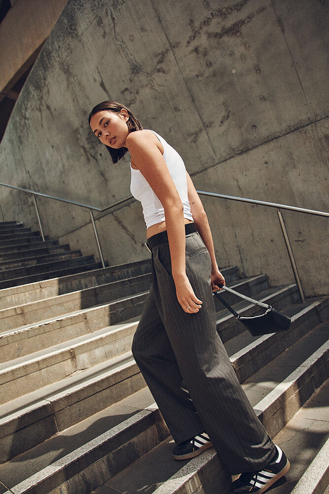 Woman in a white tank top and grey slacks walking up concrete stairs, holding a black purse, in an urban setting, shot from below.