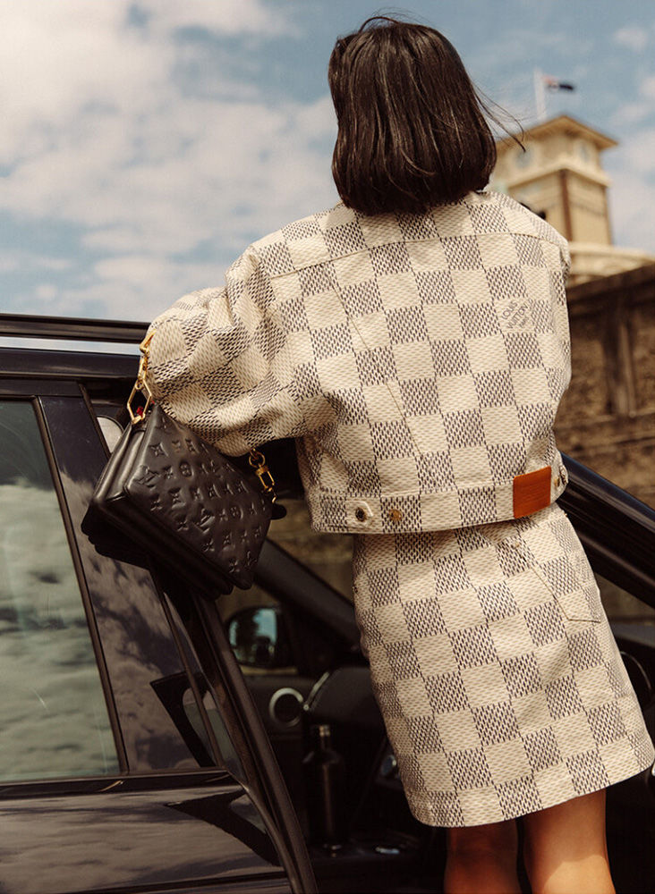 Woman in checkerboard jacket and skirt leaning against a car door holding a black handbag photographed by Dan Roberts