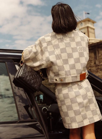 Woman in checkerboard jacket and skirt leaning against a car door holding a black handbag photographed by Dan Roberts