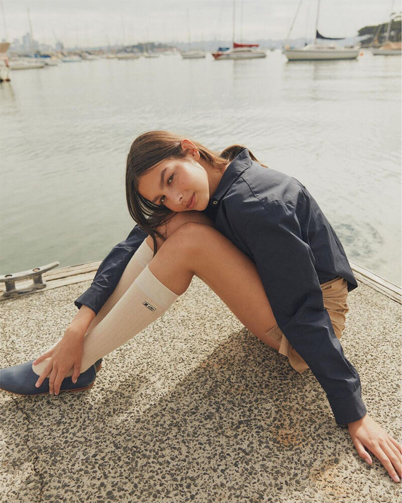 A young woman poses while sitting on a dock in front of sailboats, wearing a navy jacket, tan shorts, knee-high socks, and blue shoes.