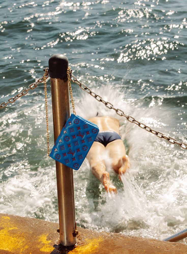 Low-angle shot of person swimming in wavy sea with blue designer bag hanging from rail, creating splash; sunny summer day.