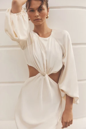 A woman poses in a white dress with cut-out details in a close-up shot against a textured white wall, with soft lighting.