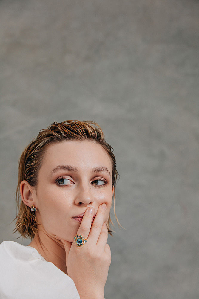 A woman with wet blonde hair and fair skin poses in front of a grey background wearing a white shirt and jewelry on her hand and ear.