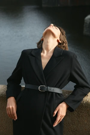 Woman in a black belted coat leaning back against a stone wall outdoors in Sydney