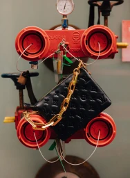Black leather quilted handbag with a chunky gold chain strap hung against industrial red valves and pipes.