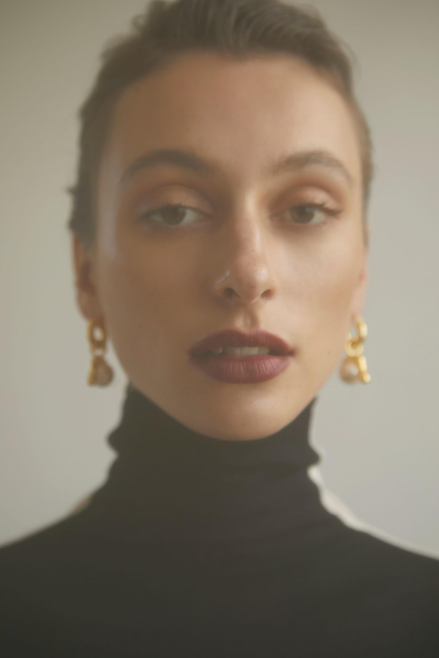 Close-up portrait of an adult female wearing gold earrings and a black turtleneck against a neutral background in soft studio light.