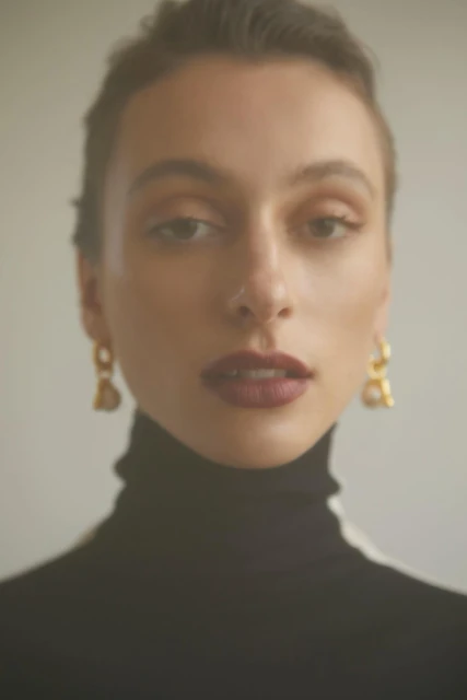 Close-up portrait of an adult female wearing gold earrings and a black turtleneck against a neutral background in soft studio light.