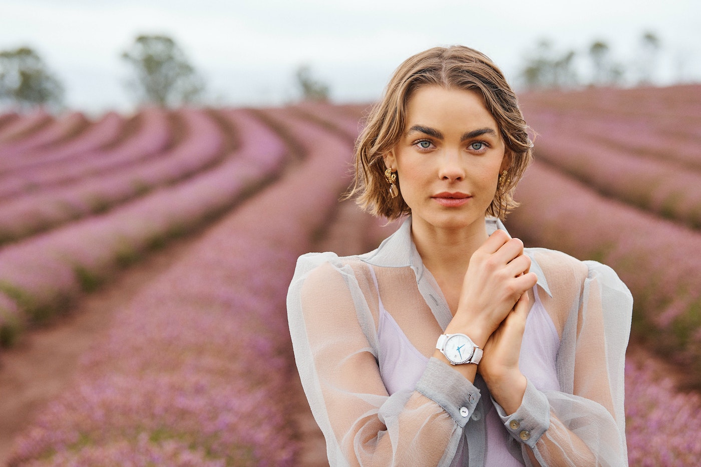 Portrait of a woman wearing a sheer blouse and wristwatch, posing in a lavender field on an overcast day with soft lighting.