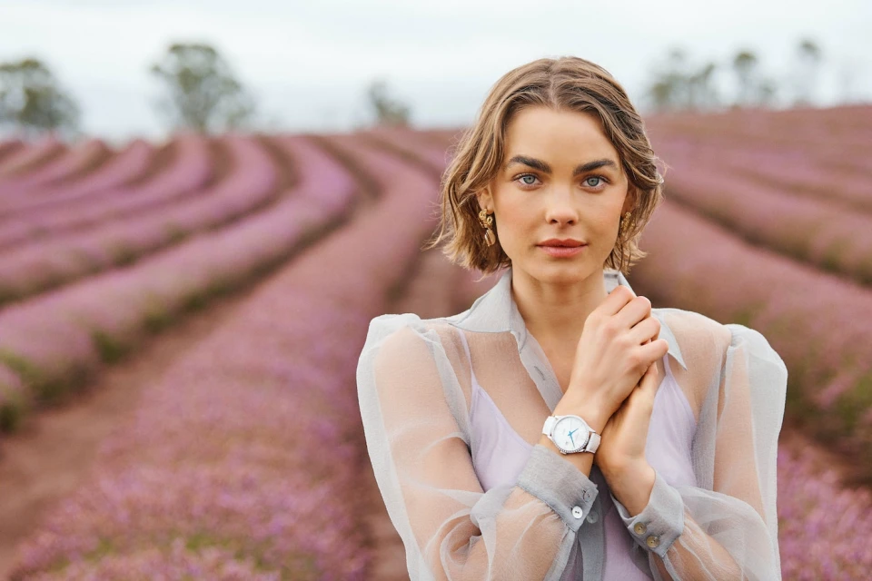 Portrait of a woman wearing a sheer blouse and wristwatch, posing in a lavender field on an overcast day with soft lighting.