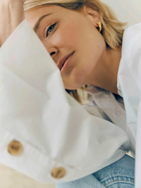 Close-up portrait of a blonde woman in a white collared shirt, featuring her blue eyes, golden hoop earrings, and fair skin.