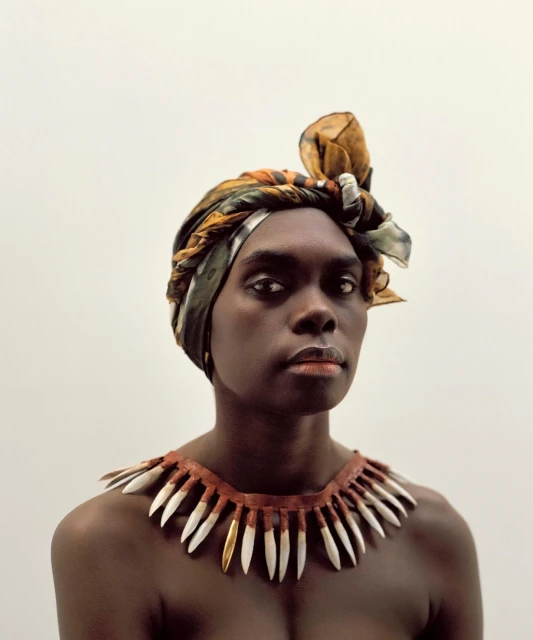 Striking portrait of a woman with dark skin wearing a patterned headscarf and a tooth necklace against a plain white backdrop.