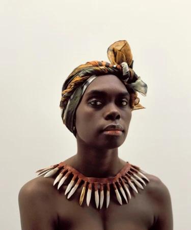 Striking portrait of a woman with dark skin wearing a patterned headscarf and a tooth necklace against a plain white backdrop.