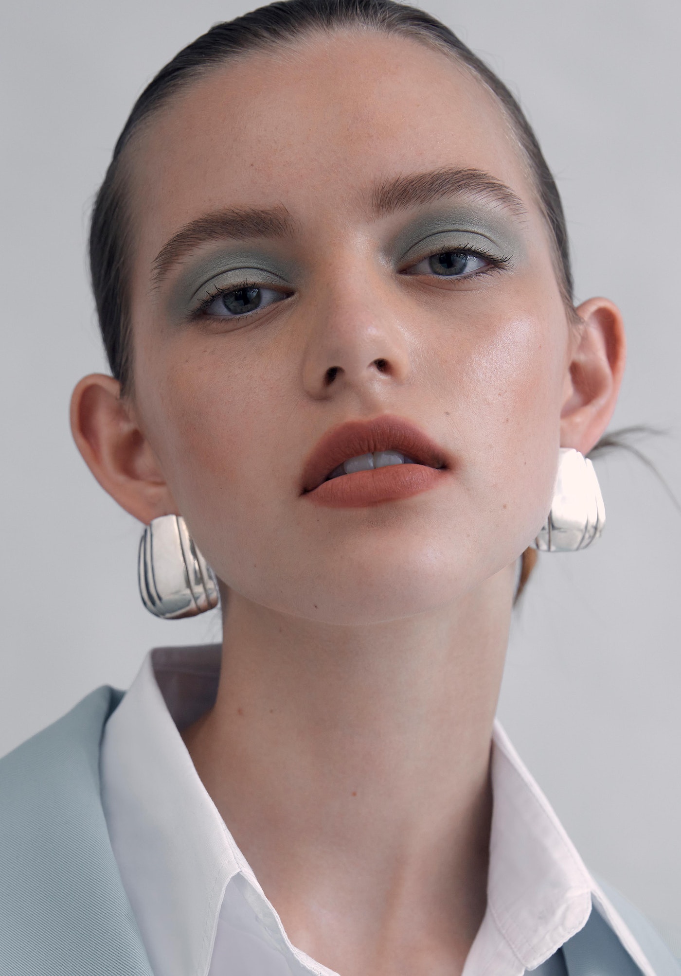 Close-up of a young woman with light skin and green eyeshadow wearing square silver earrings, a white collared shirt and a light blue blazer.