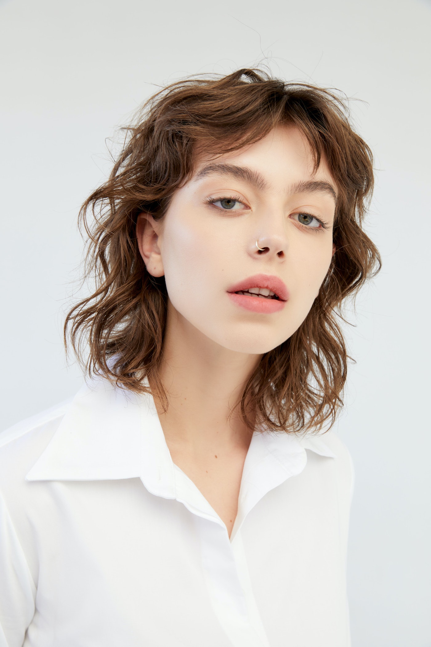 Portrait of a young woman with brown curly hair and wearing a white button-down shirt against a neutral background in soft, diffused lighting.