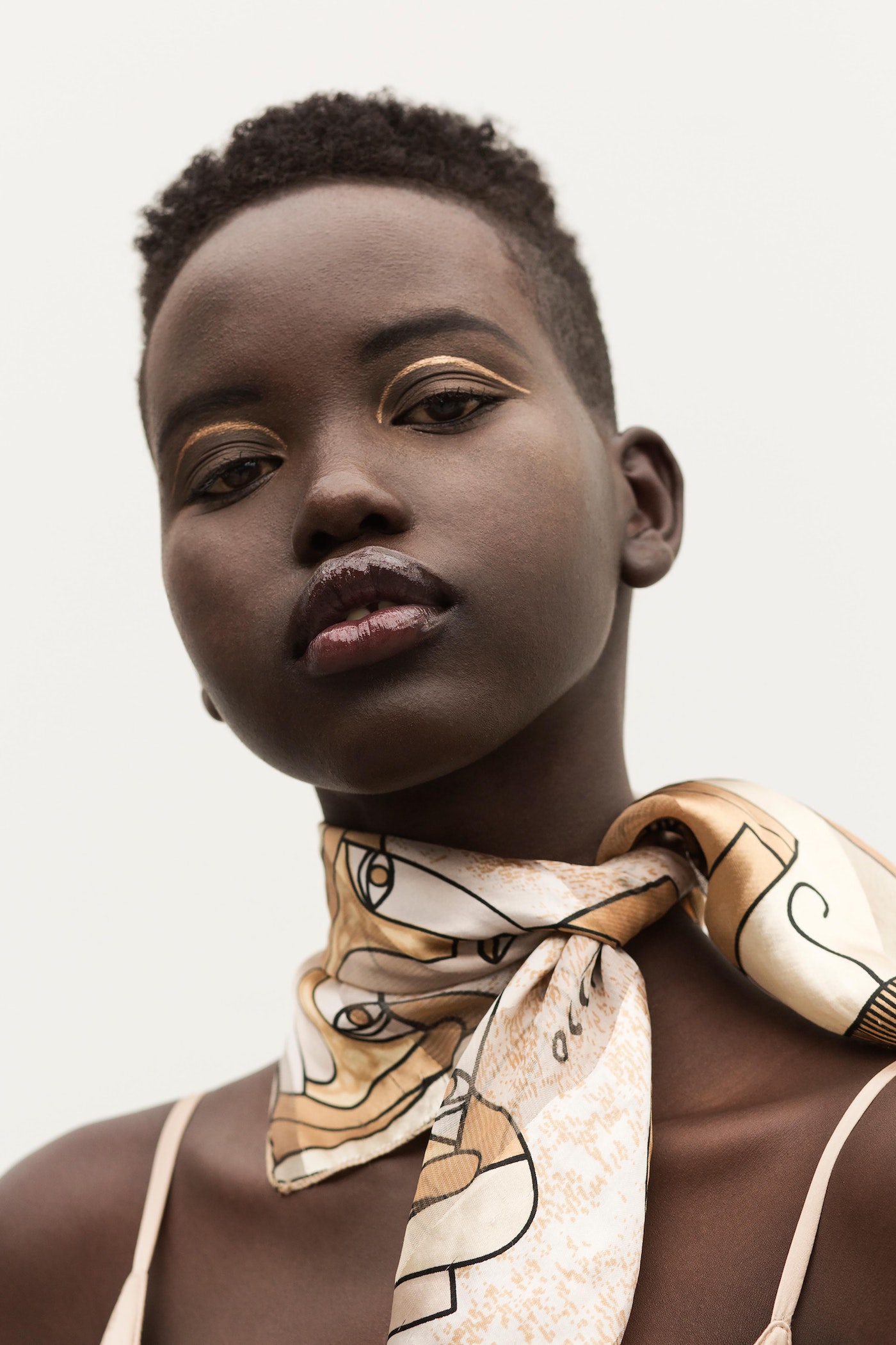 Close-up portrait of a beautiful young African-descent woman with gold eyeshadow and a patterned scarf against a white background.