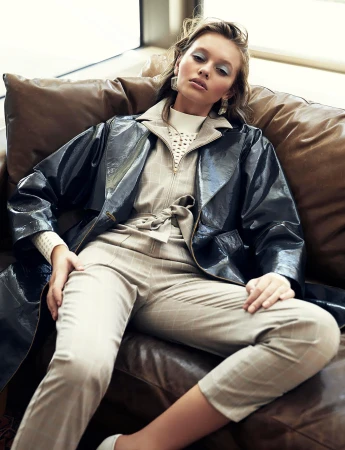 Model seated on a brown leather sofa wearing a belted beige jumpsuit and a dark glossy trench coat