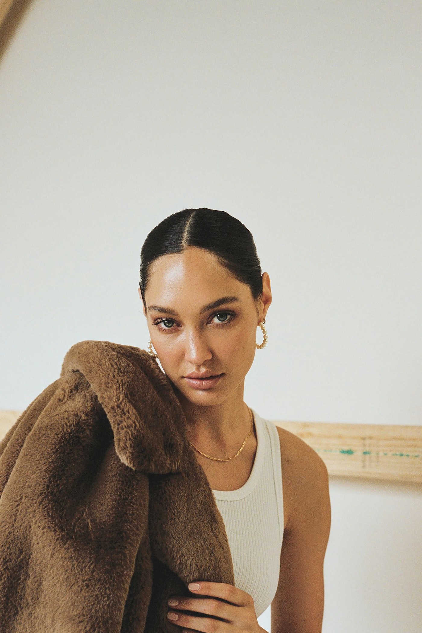 Woman in a tank top with a brown fur coat draped over her shoulder, posing in a studio with bright, neutral lighting and clear focus.