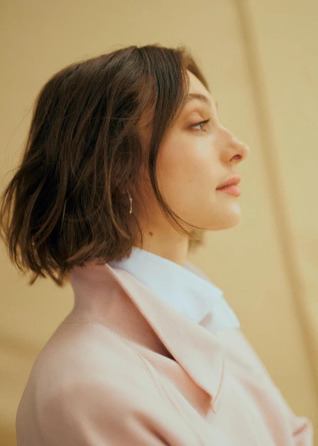 Profile of a young adult woman with short brown hair wearing a light pink blazer and white collared shirt against a neutral background.