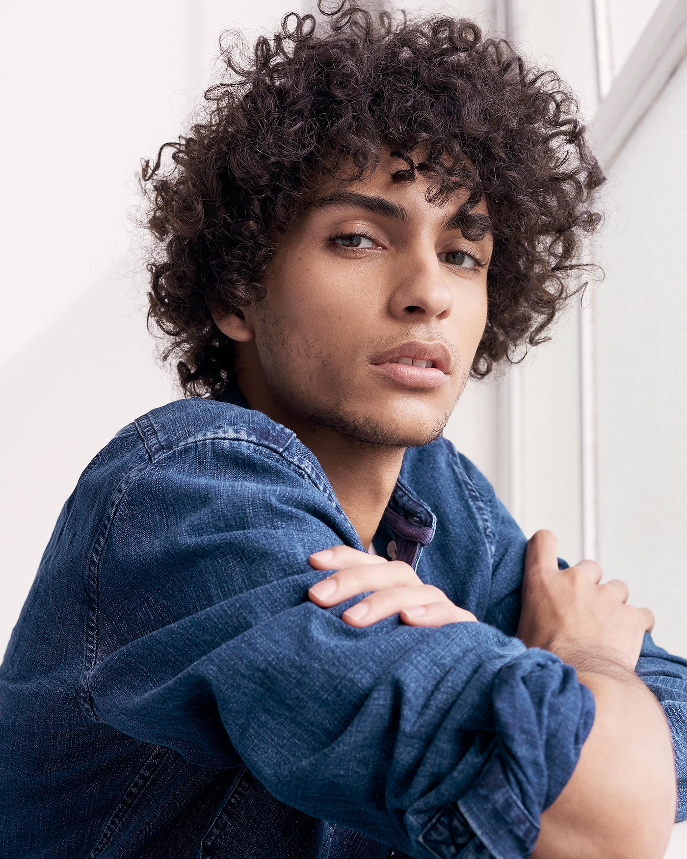 Close-up of a young adult male with curly hair, wearing a denim shirt, posing indoors with arms folded and looking directly at the camera.