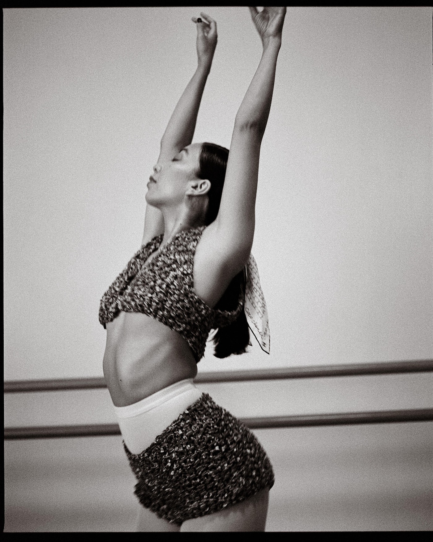 Black and white photo of an Asian woman in knitwear posing with arms raised in a dance studio with barre in the background.