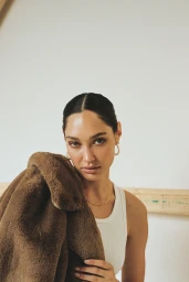 Portrait of a woman holding a brown faux fur jacket wearing a white tank top with slicked-back dark hair
