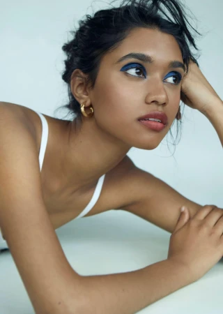 Close-up portrait of a south asian woman with bold blue eyeshadow and gold hoop earrings posing against a white background