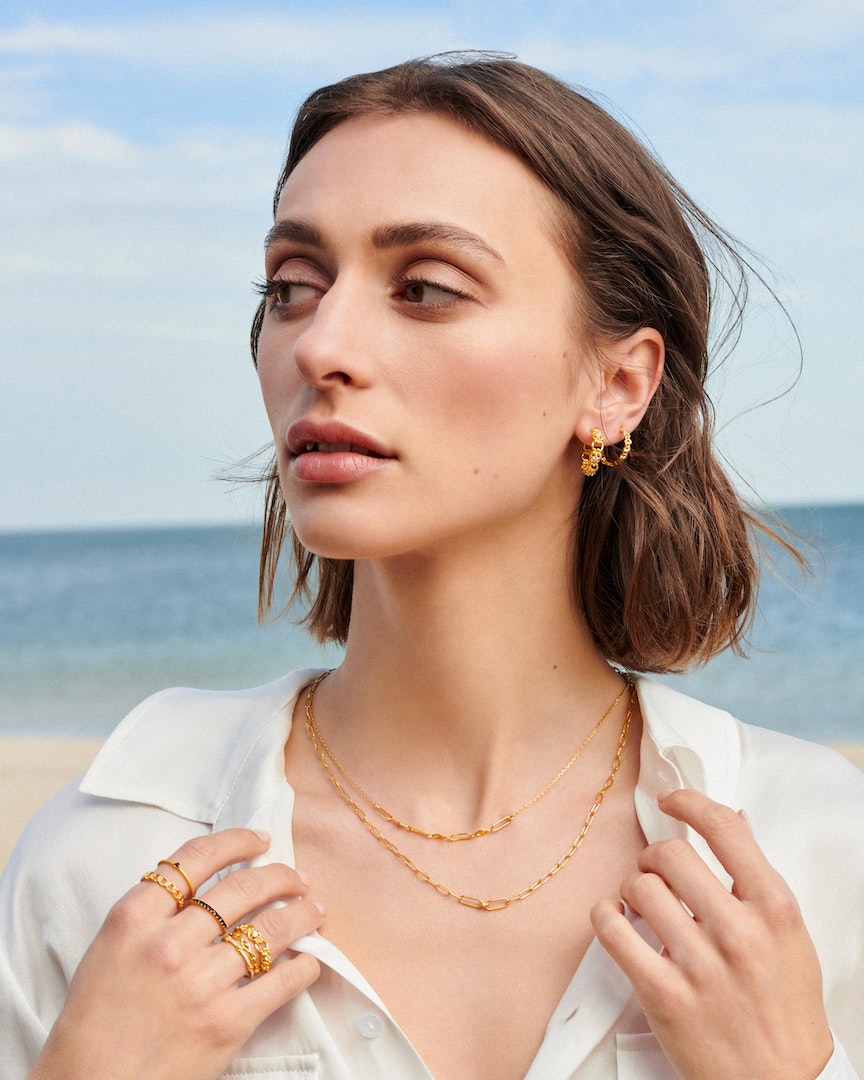 Close-up of a brunette woman wearing gold jewelry, including a necklace, earrings, and rings, posing outdoors near a beach.