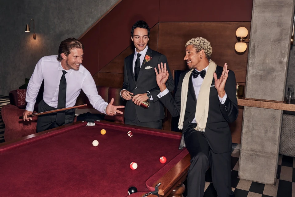 Three men in formal attire playing pool. One man is aiming with a cue, another is holding a bottle, and the third is seated.