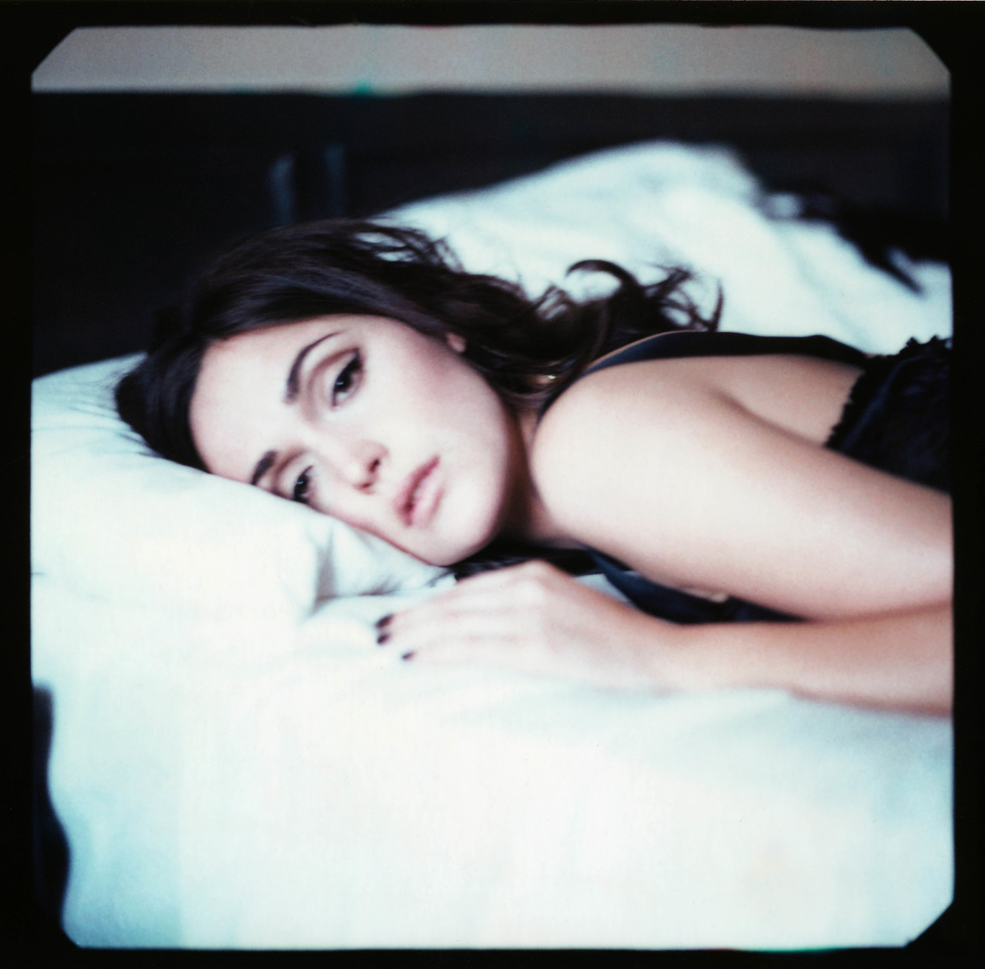 Brunette woman in a black tank top poses while lying on a white pillow and bedsheet in a bedroom setting with dark wood furniture.