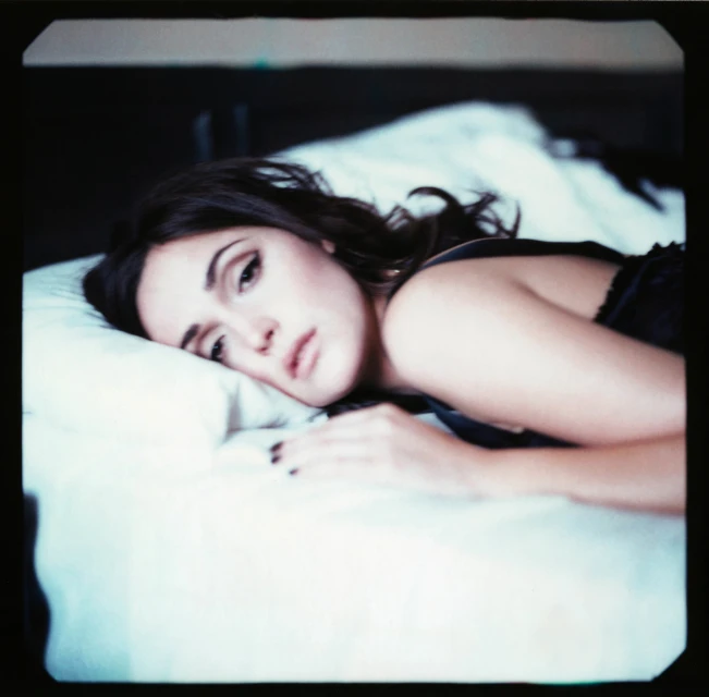 Brunette woman in a black tank top poses while lying on a white pillow and bedsheet in a bedroom setting with dark wood furniture.