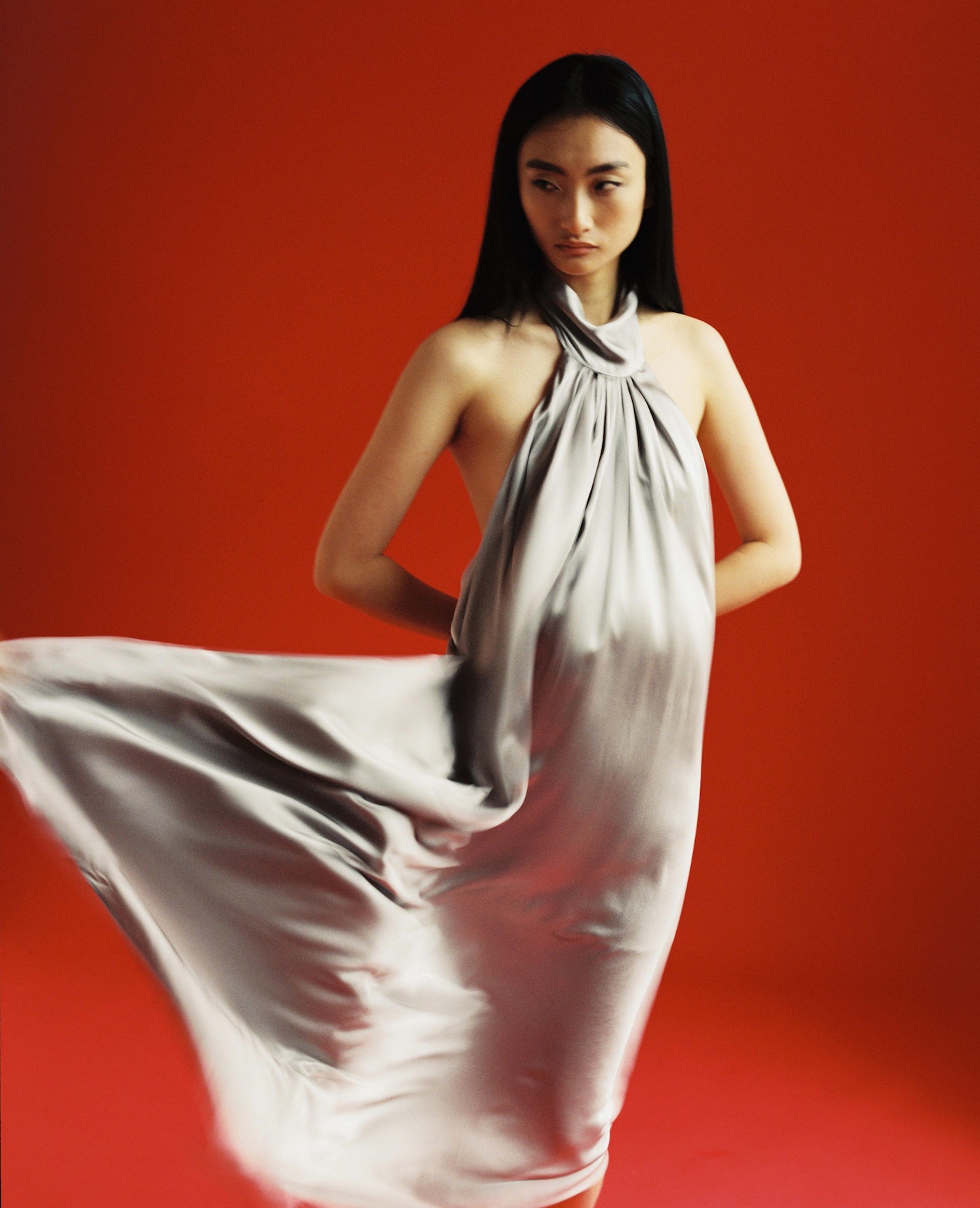 Asian model in a silver dress posing against a red background in a studio setting, fashion editorial photography with simple background.