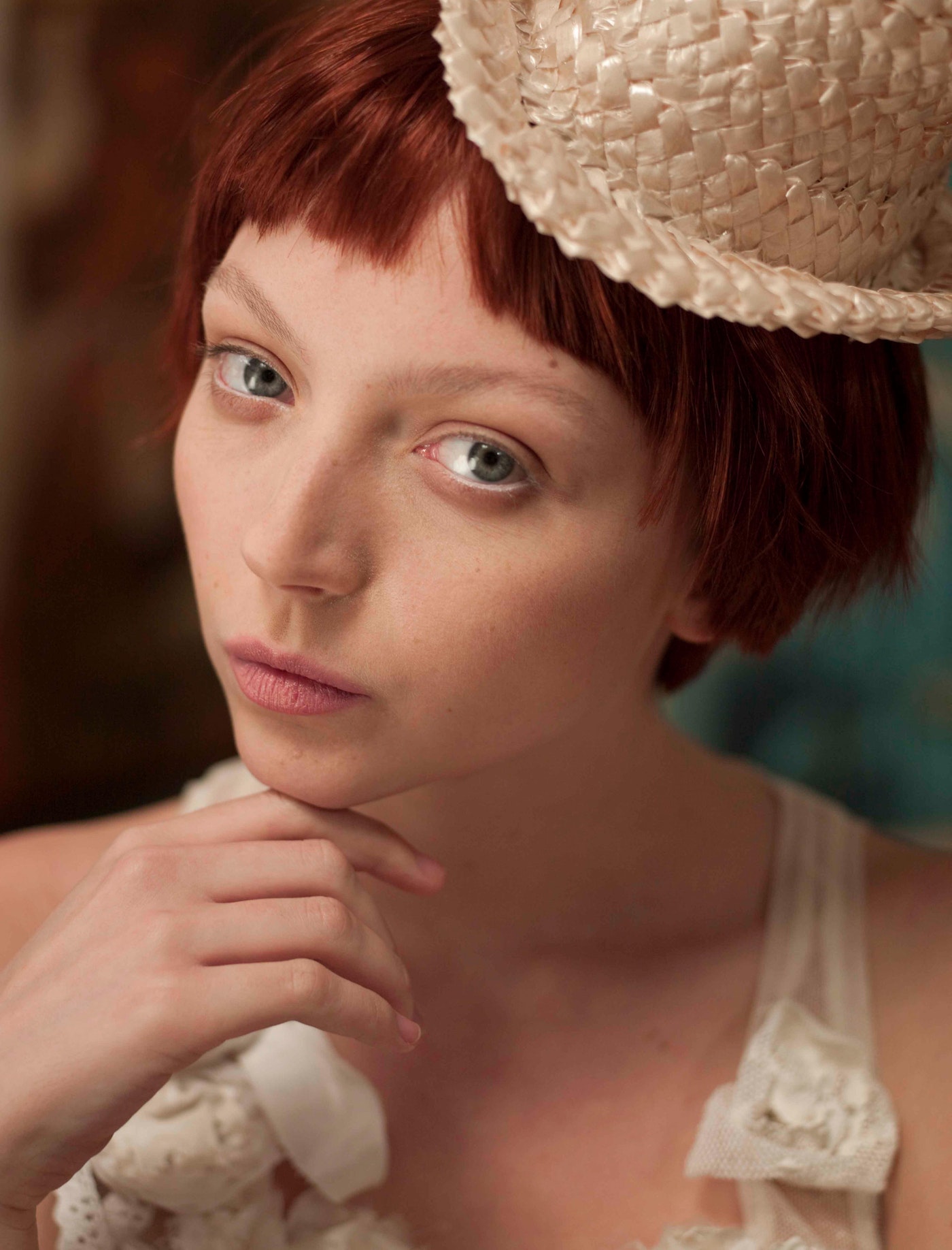 Close-up studio portrait of a red-haired woman in a straw hat with her hand under her chin, wearing a lacy white top, and looking at the camera.