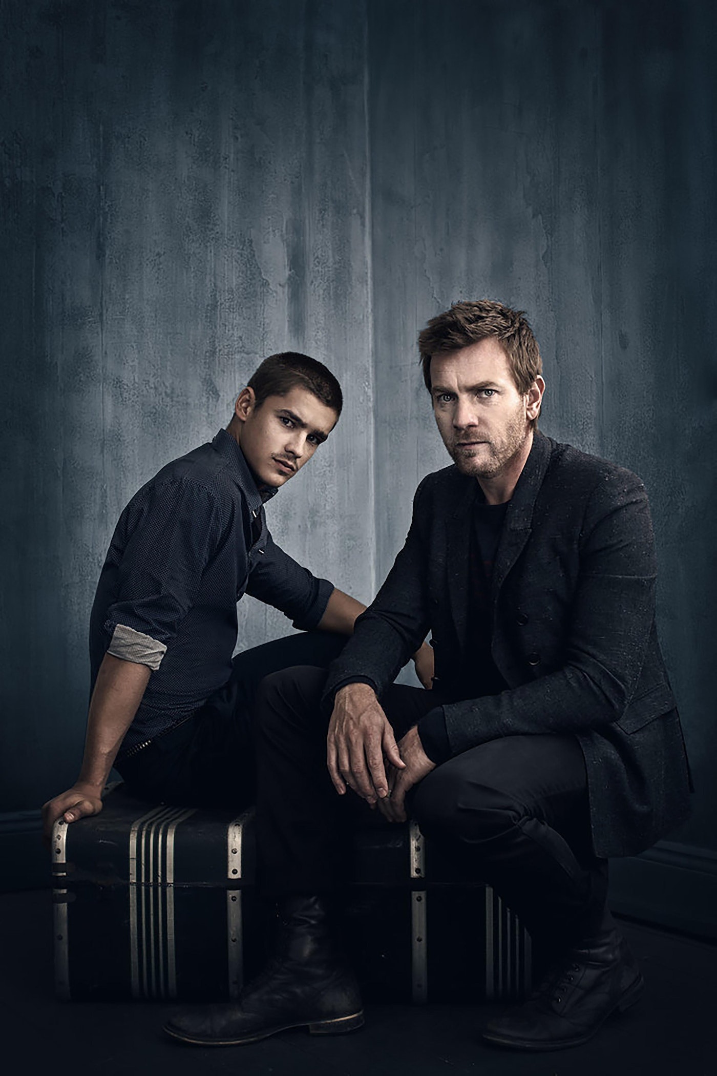 Two adult men sit on a trunk wearing dark clothing in a studio with a grey textured background, posing for a portrait.