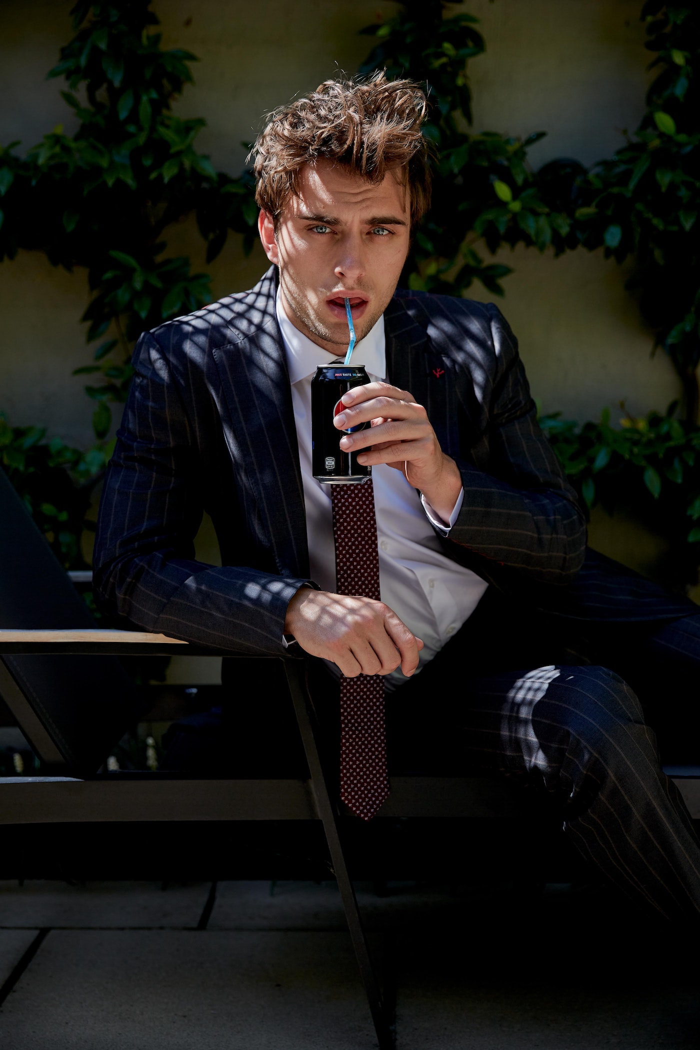 A young adult male in a suit and tie sits and drinks from a can with a straw in an outdoor setting with green foliage in the background.