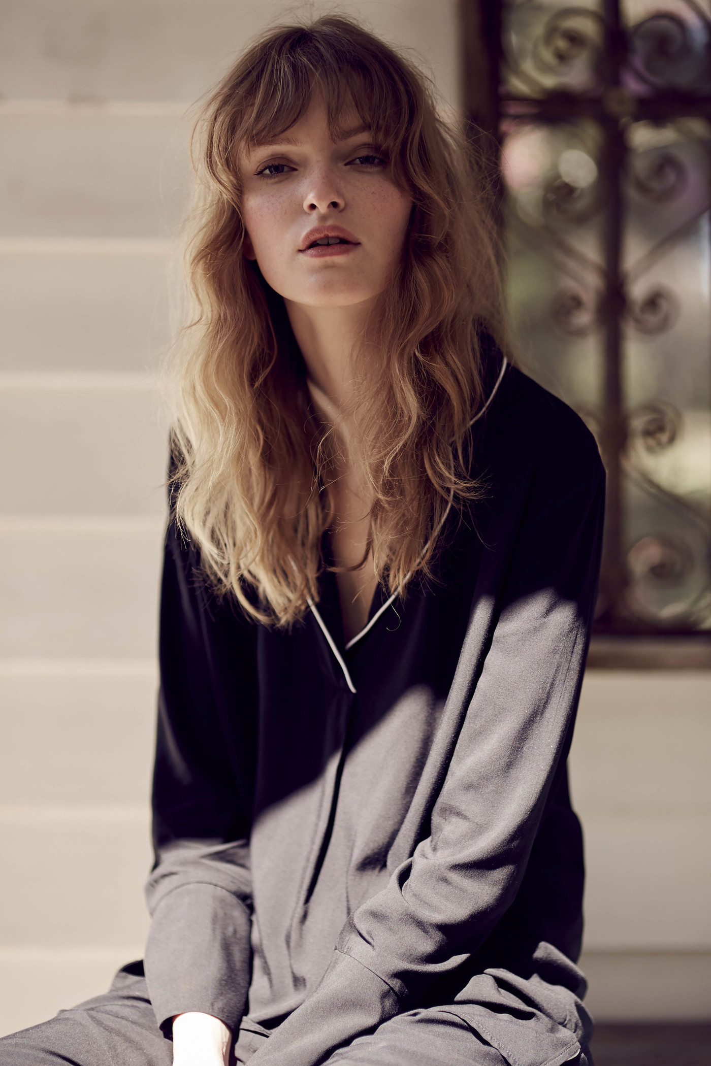 Blonde woman in black lounge wear sits in front of a window with patterned ironwork with sunlight on her face and wavy long hair.