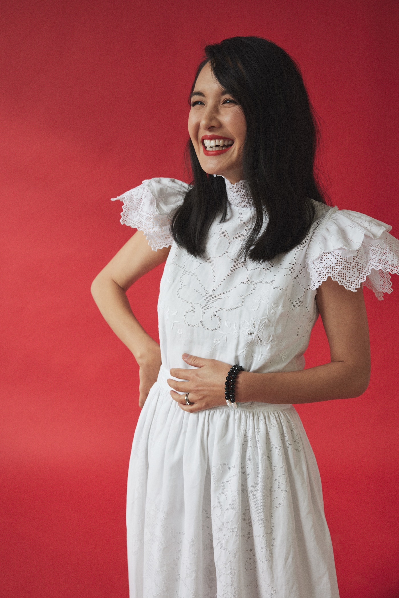 Smiling woman in a white dress poses against a red background. She has dark hair and red lipstick and is looking off to the side.